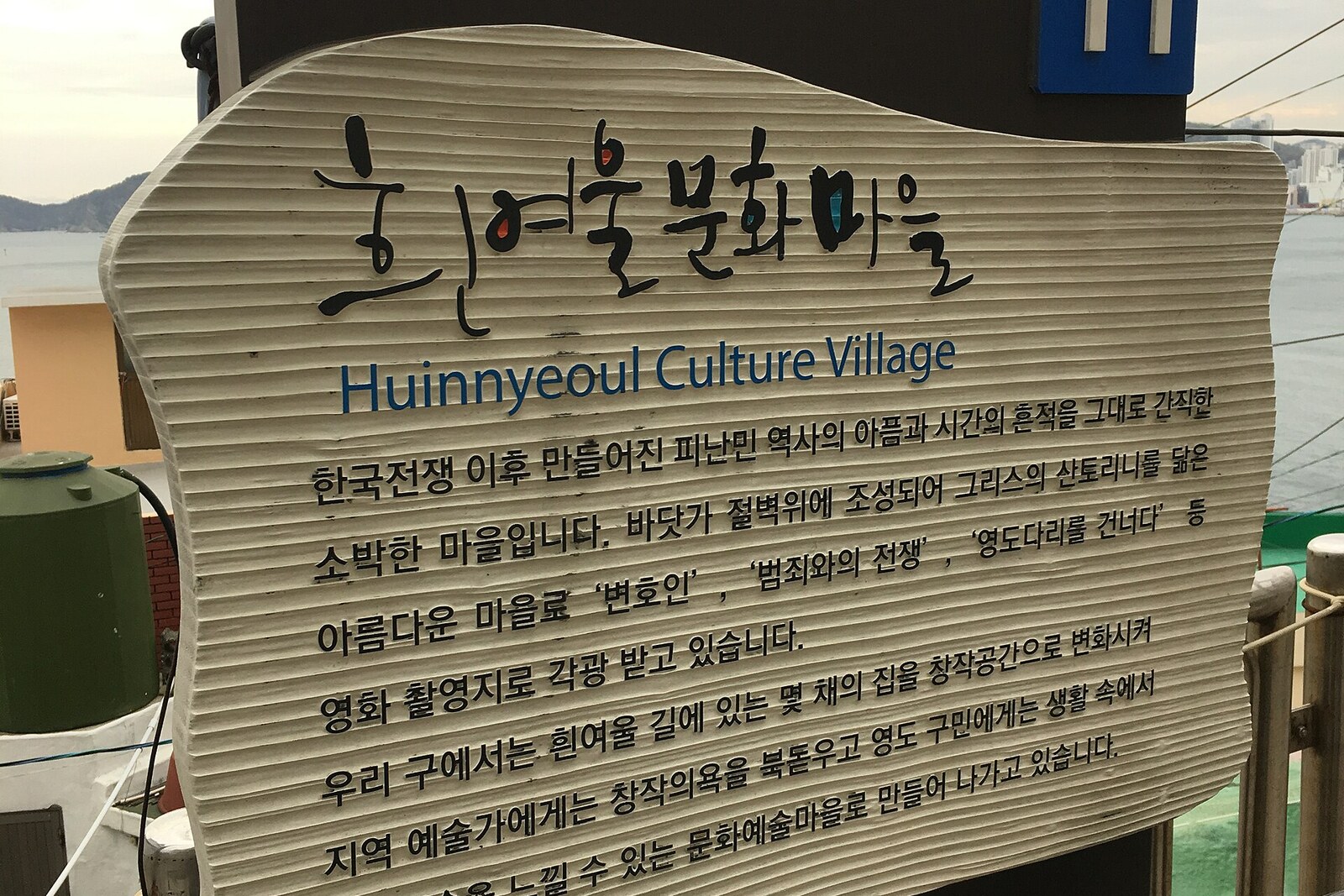 A wooden information board at the entrance to Huinnyeoul cultural village, with the sea in the background.