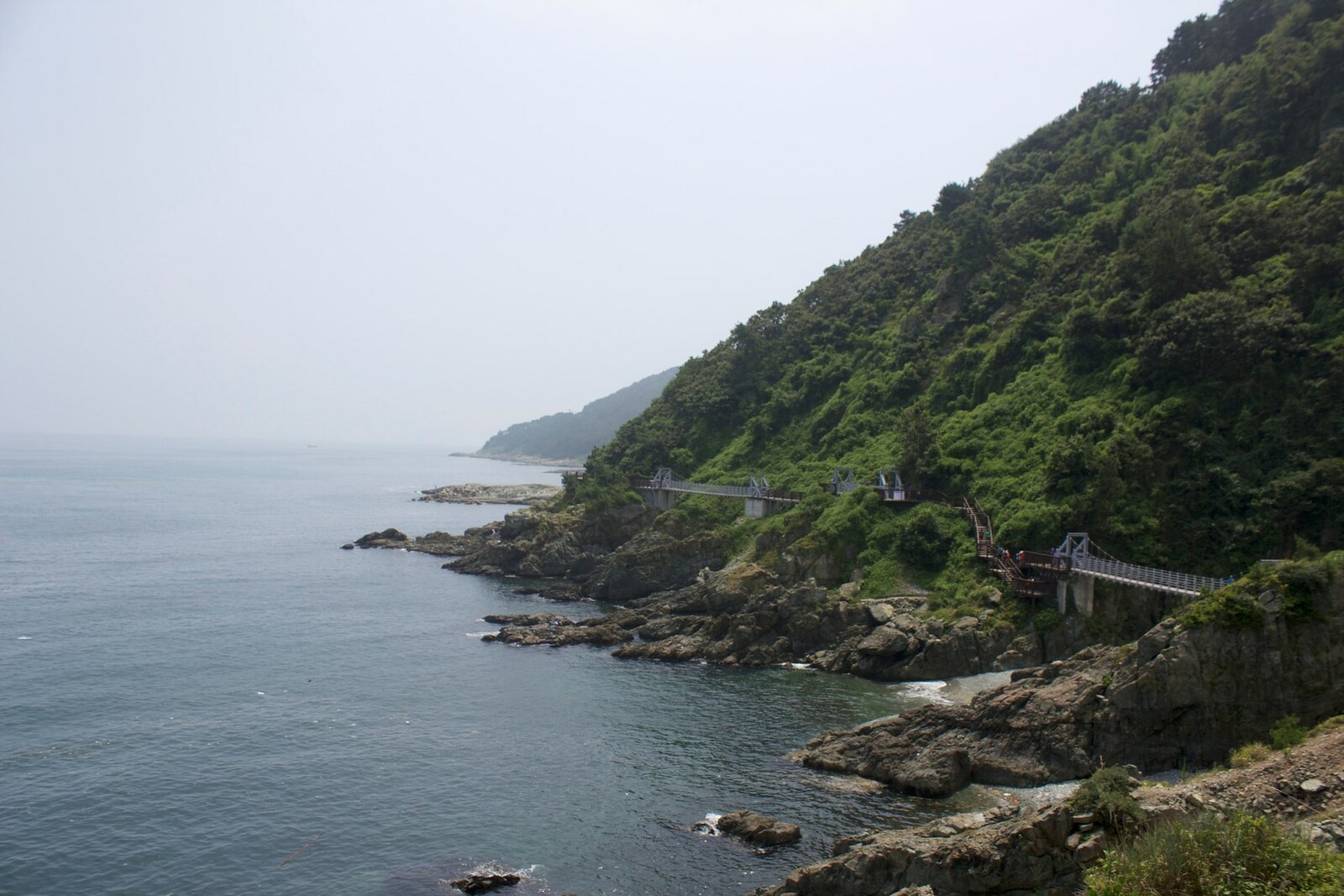 A trail along cliffs with a suspension bridge above coastal rocks.