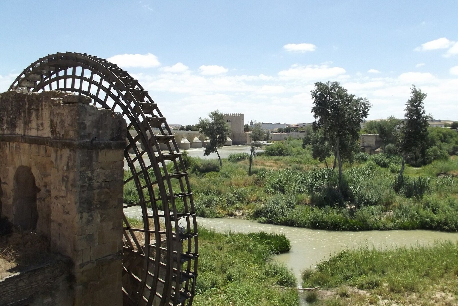 The wooden Albolafia wheel beside a stone structure on the bank of the Guadalquivir.