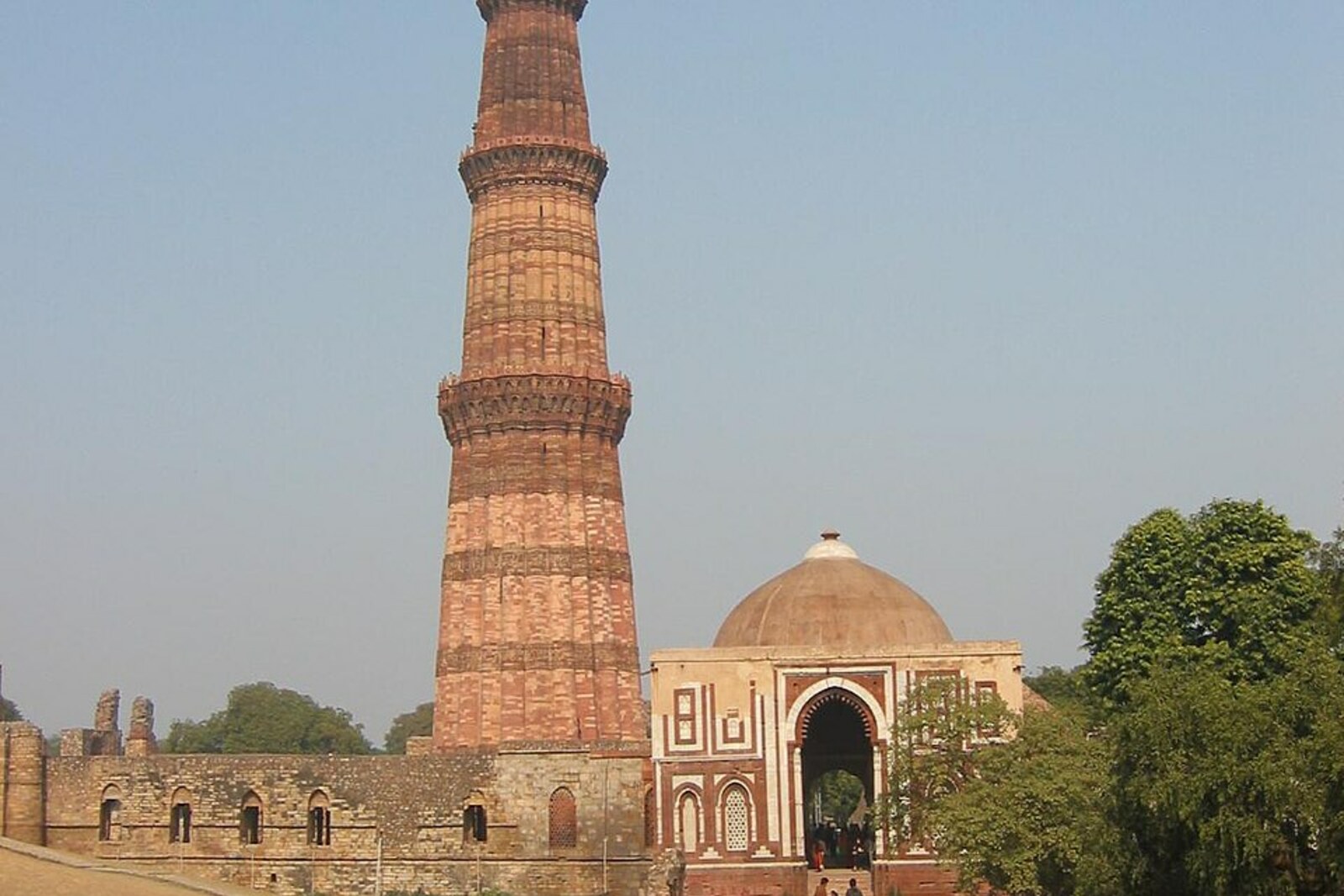 Qutb Minar rises above green trees against a clear sky.