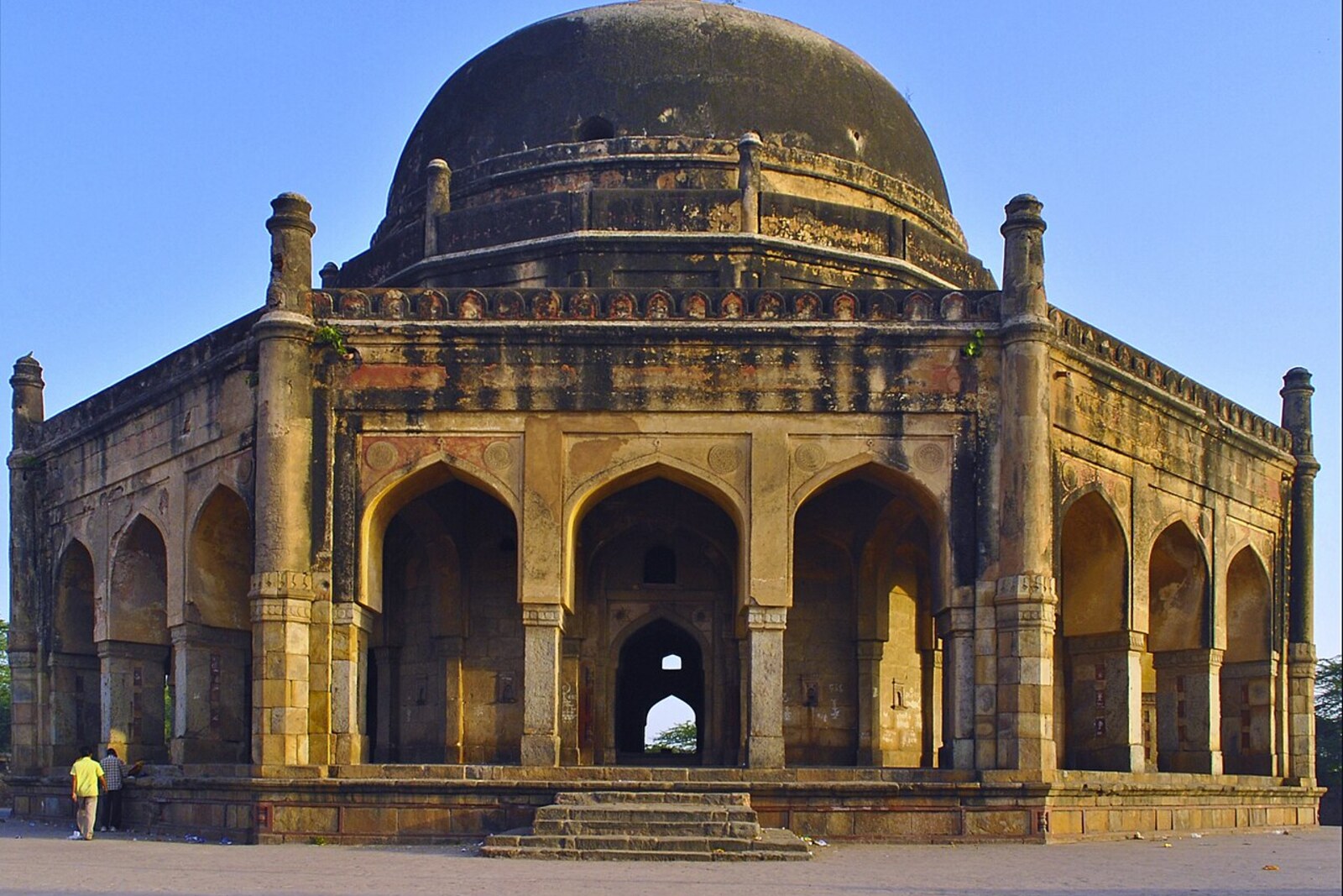 An octagonal tomb with a dome and arches on a sunlit slope.