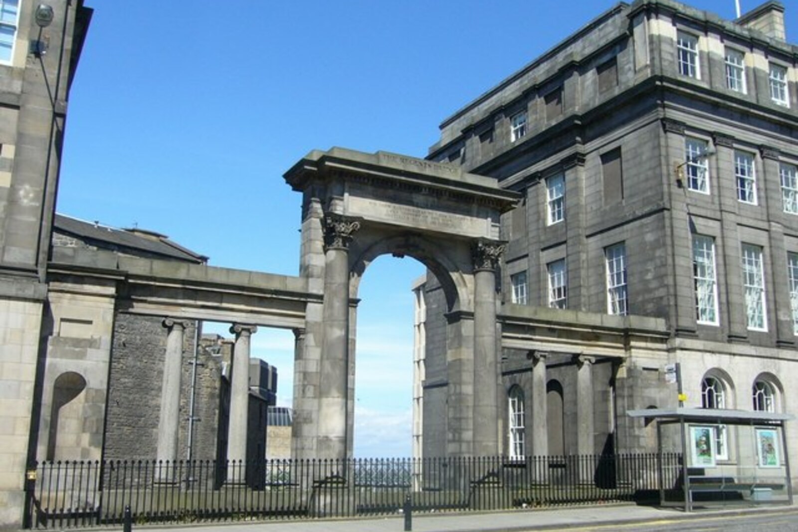 A stone bridge with arches and Doric columns spans a street; the green slope of Calton Hill is visible behind it.