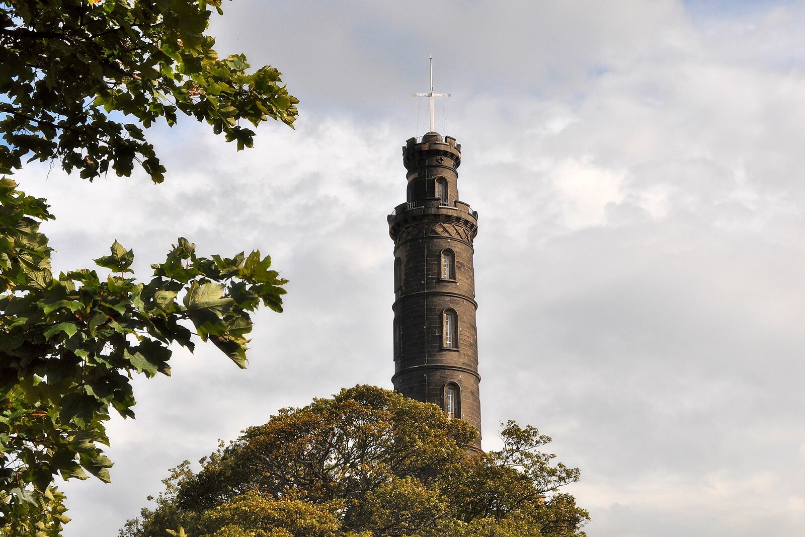 A stone tower with a balcony and a mast at its summit rises above a green hill and blue sky.
