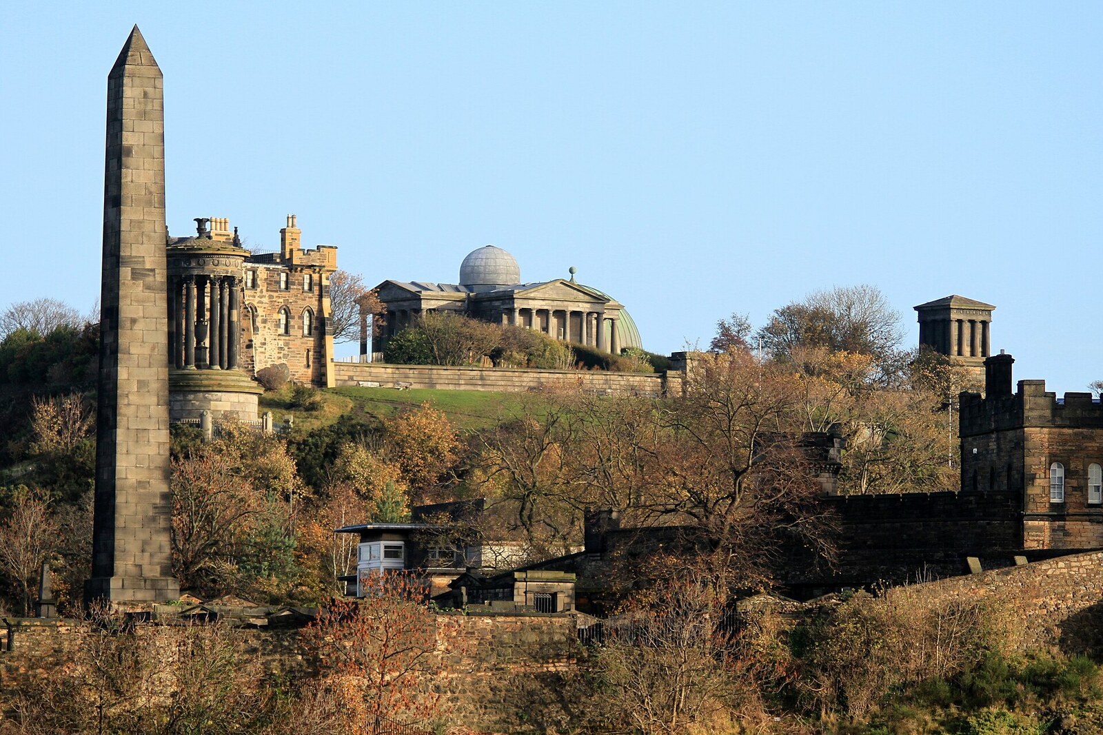 Low white-stone buildings with domes and a tower stand on the summit of a hill; green grass fills the foreground.