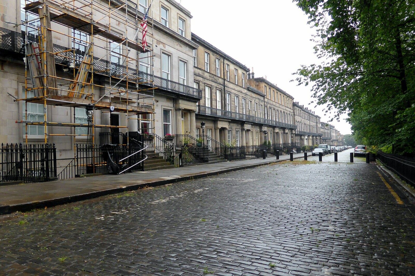 A row of stone houses with Doric columns and black railings on a quiet street; green lawns lie ahead.