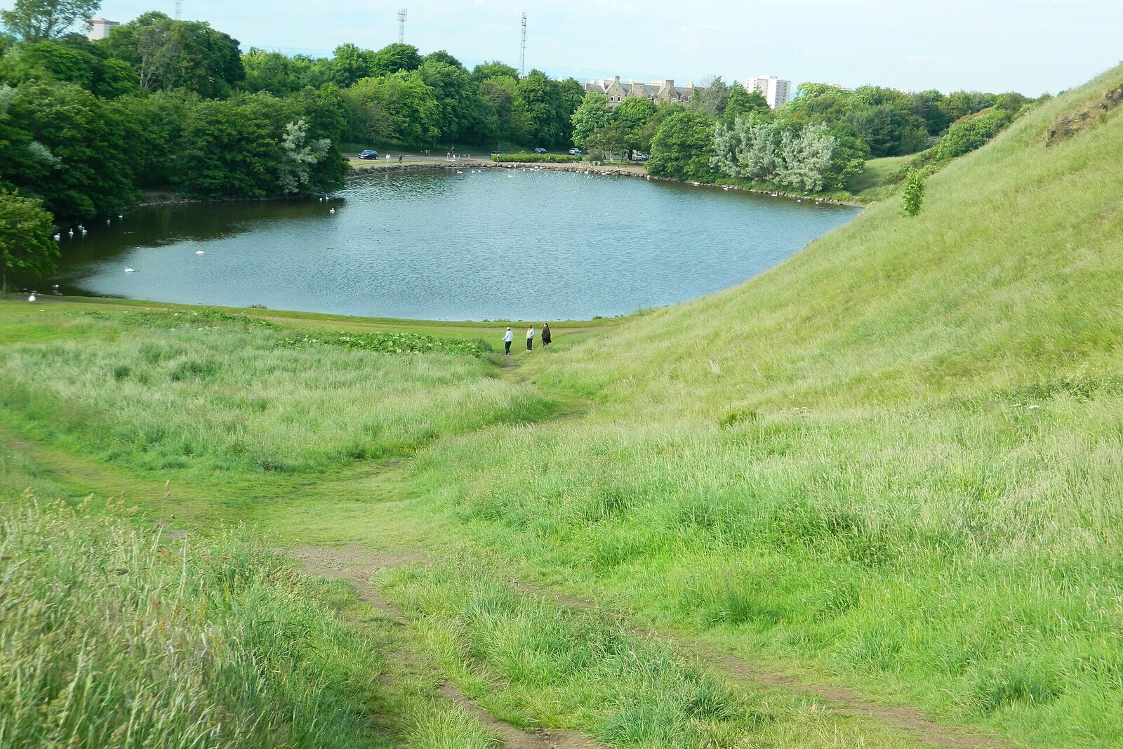 A calm loch reflecting green slopes; stone ruins are visible on top of a cliff.