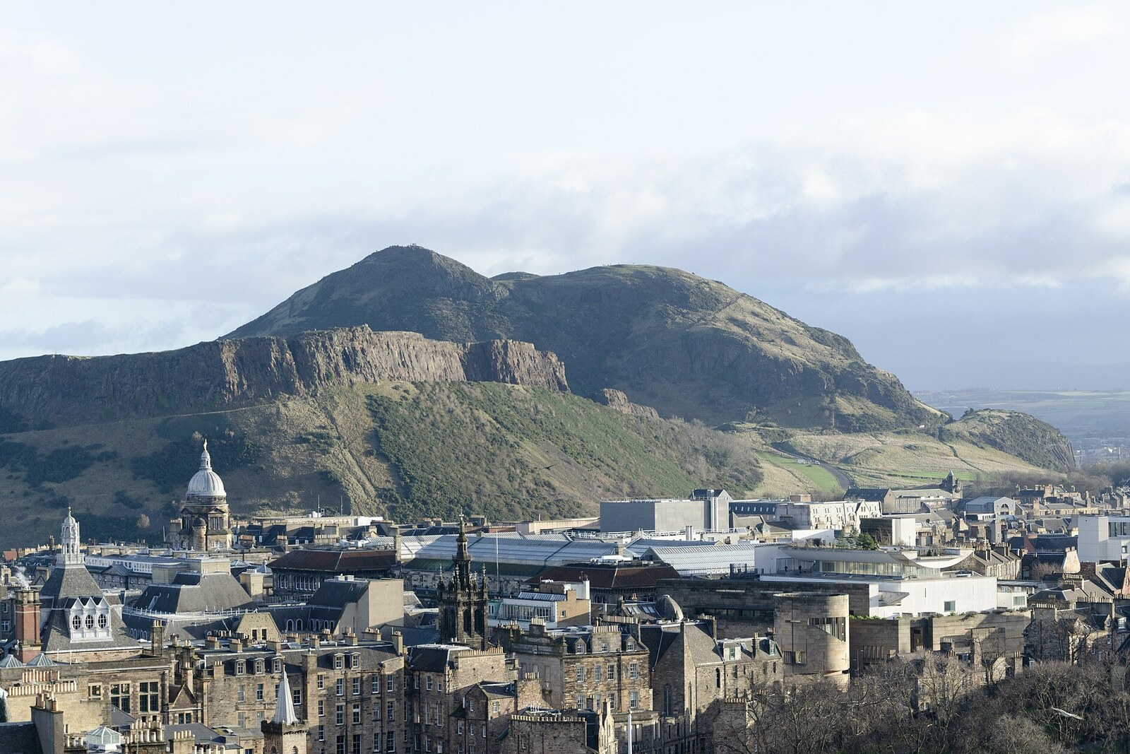 Dark rocky crags rise above green meadows; the hill of Arthur's Seat is visible in the background.