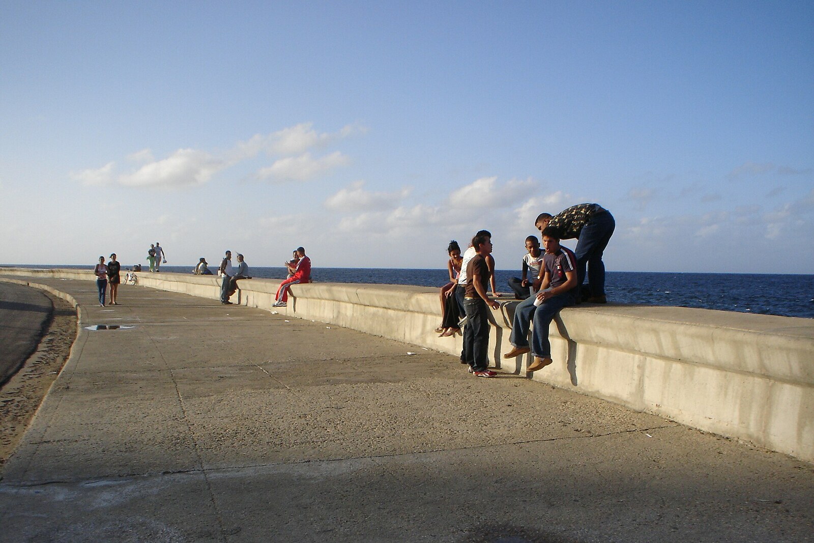 People walk along the Malecón waterfront in Havana; waves break against the wall, while colorful buildings can be seen in the background.