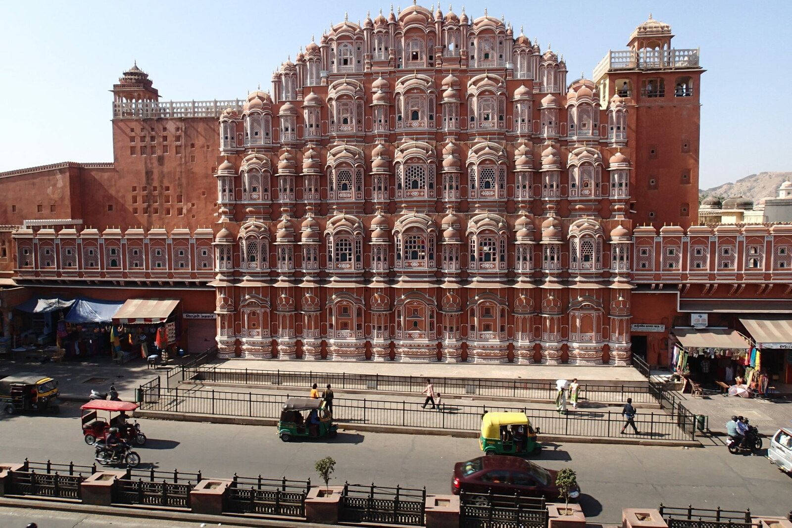 The broad Badi Chaupar square with a marble fountain and the pink facade of the Palace of Winds in the background; auto-rickshaws pass in the foreground.