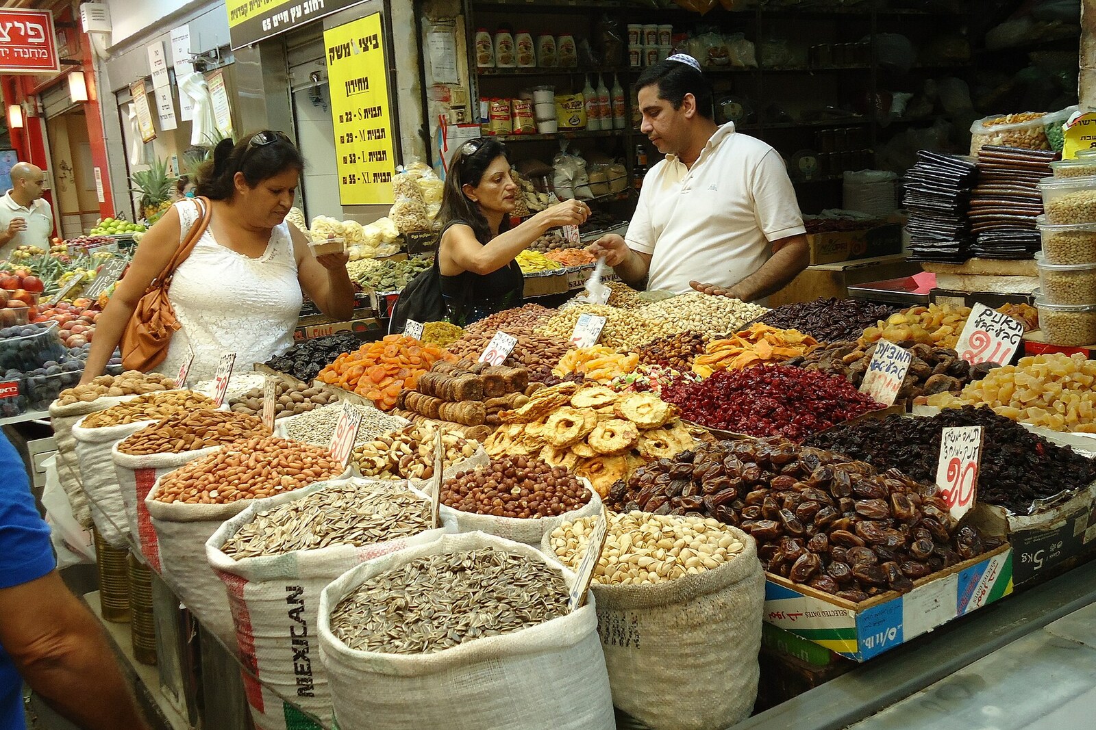A crowd of people walks between rows of fruit and vegetable stalls in Mahane Yehuda Market