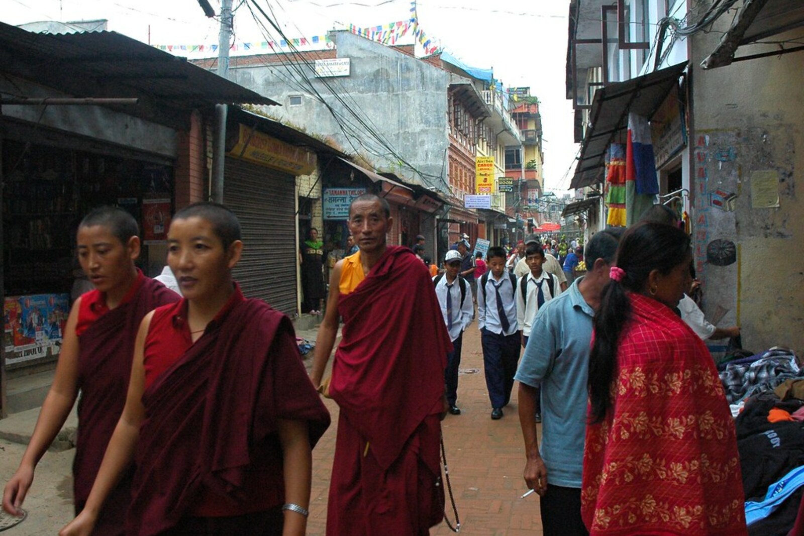 Escena callejera de Boudha en Katmandú con monjas, un monje, alumnos, tiendas y cables.