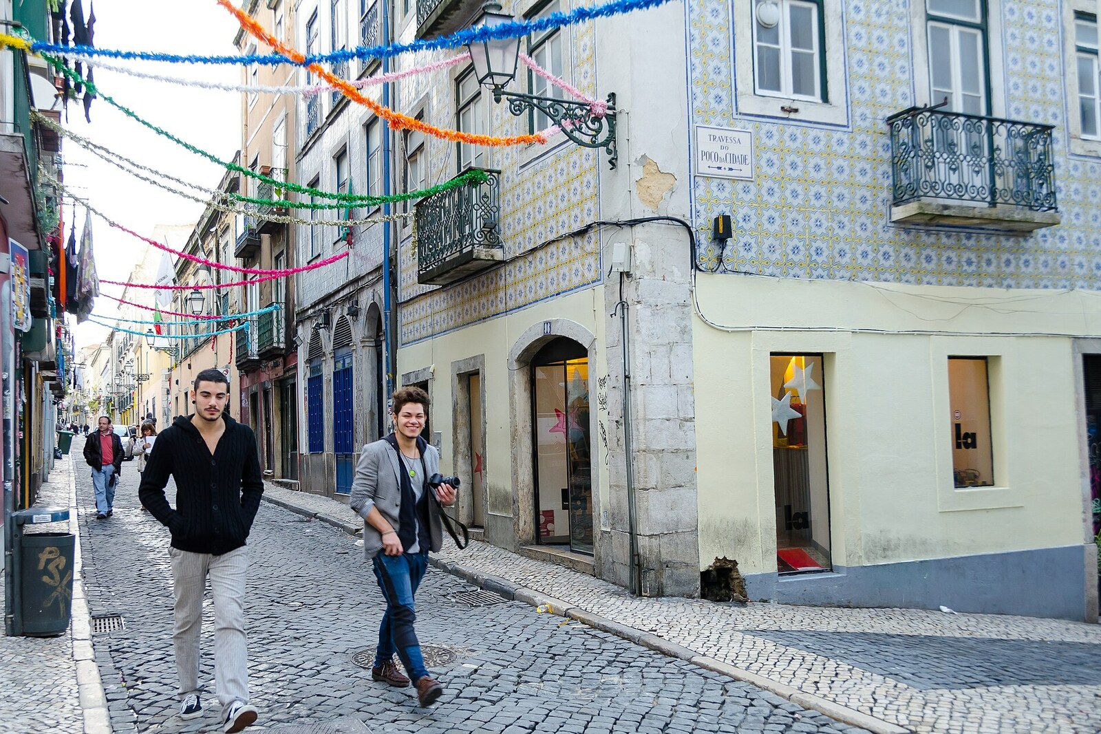 A narrow street in Lisbon with pastel-colored buildings and signs for small bars.