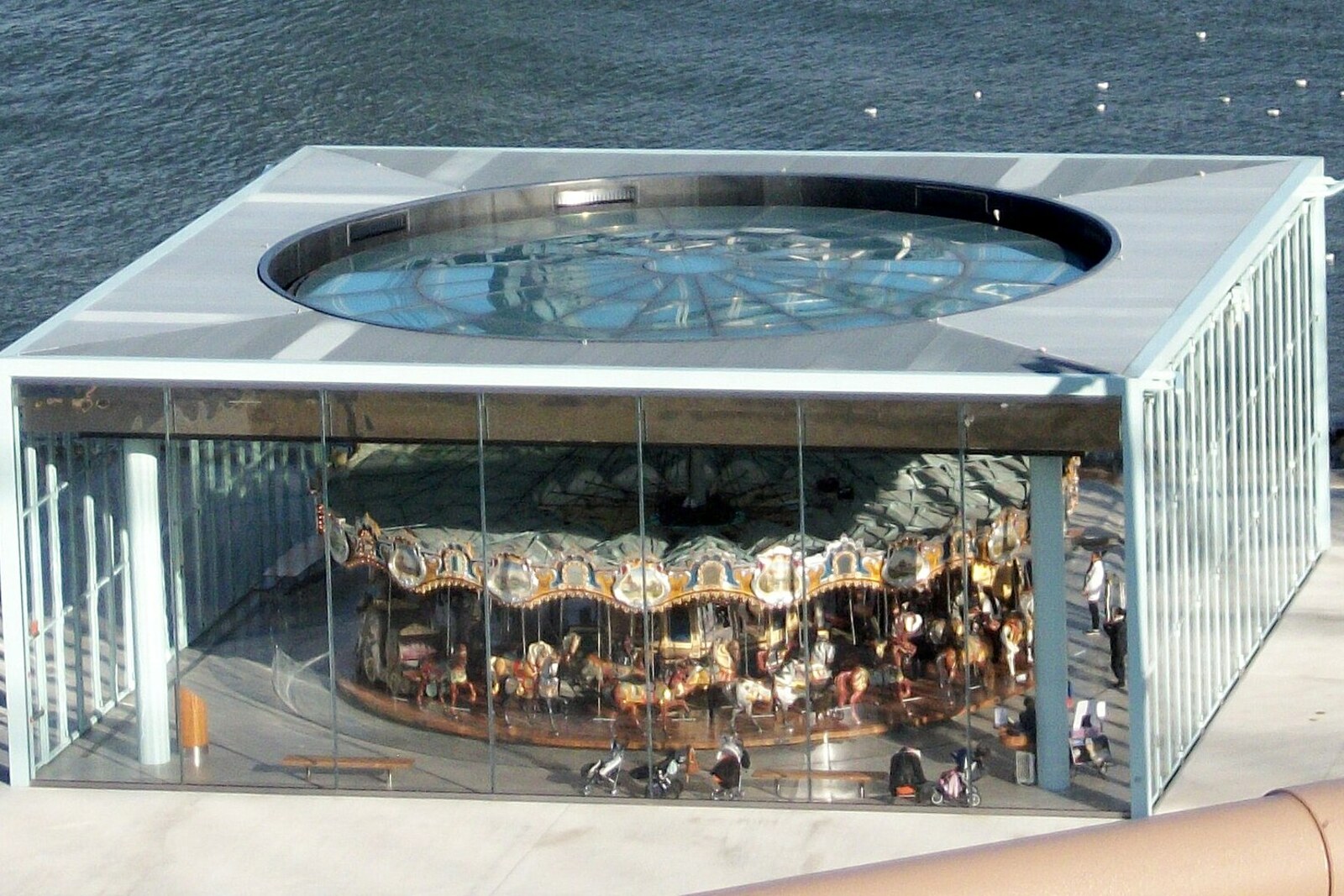 A carousel with carved horses turns inside a glass pavilion, with a bridge visible in the background