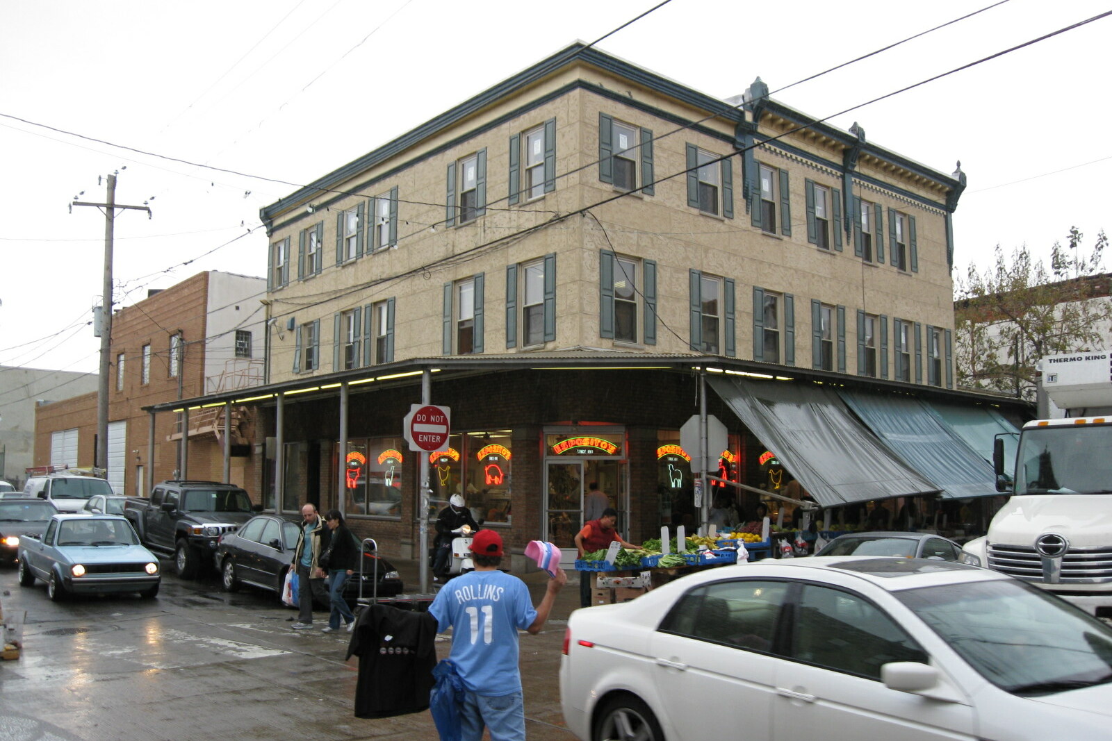 People walk through the South 9th Street Italian Market: stalls and carts stand on both sides of the street, with awnings above them.