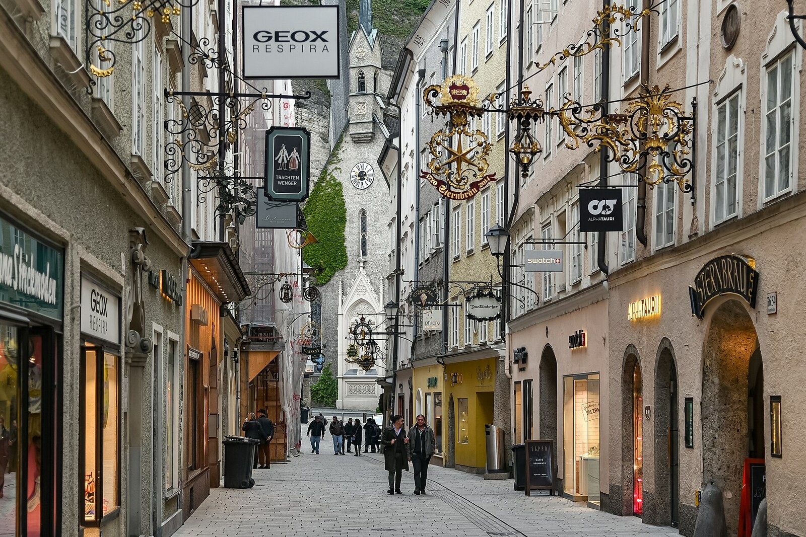 Pedestrians on a narrow street with wrought-iron signs and tall buildings