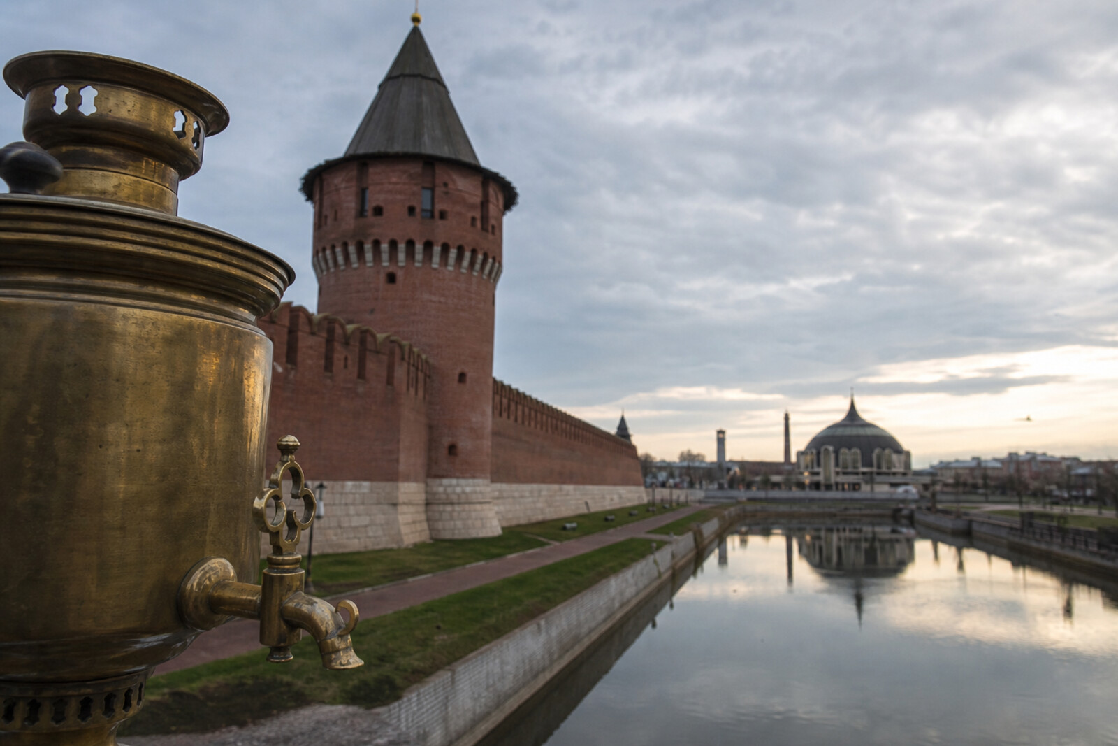 A route view of Tula: the red-brick Kremlin wall by the Upa, a brass samovar accent, and the silhouette of the helmet-shaped museum.