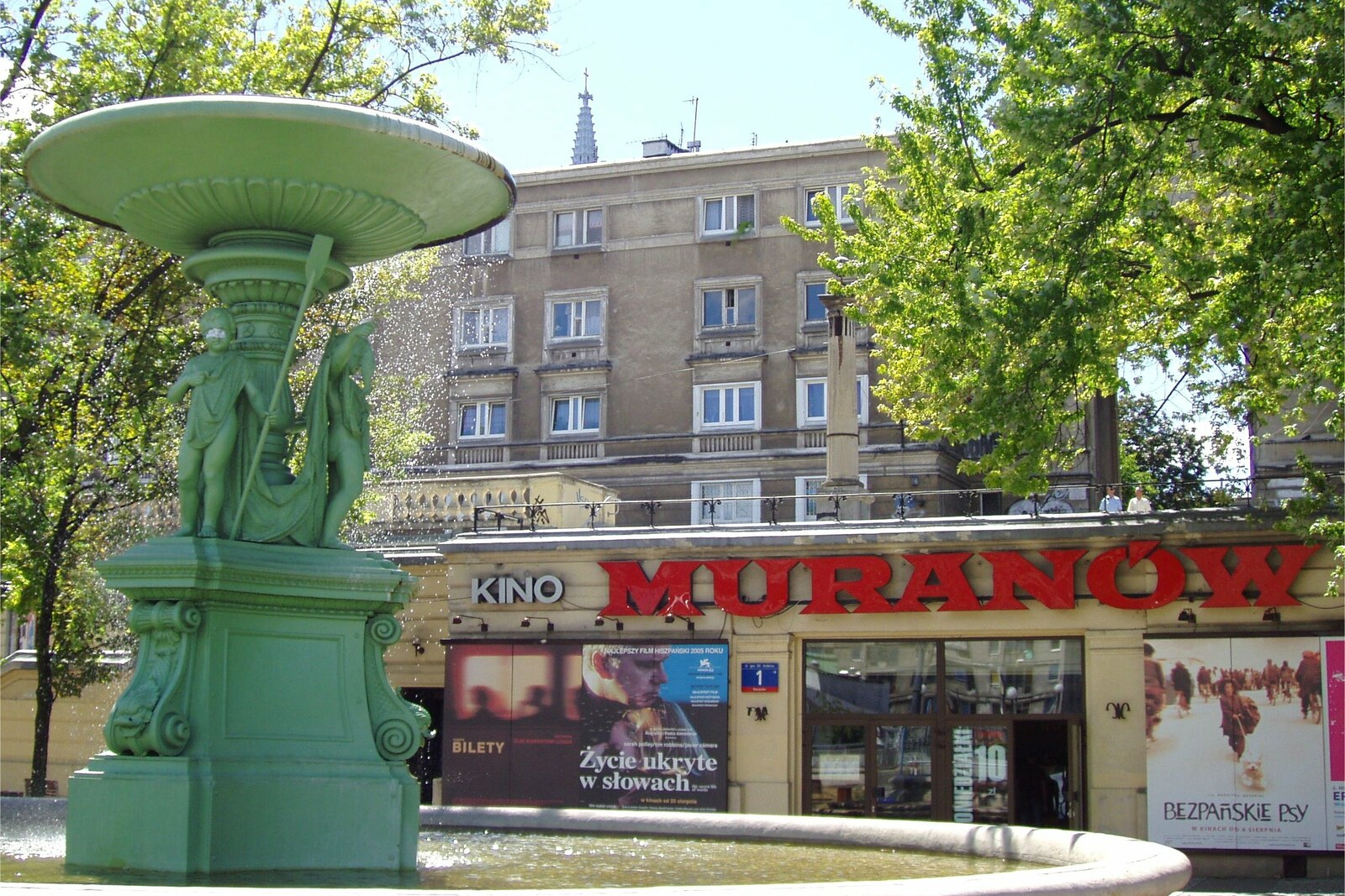 A historic fountain and the cinema facade on a quiet, tree-lined square.