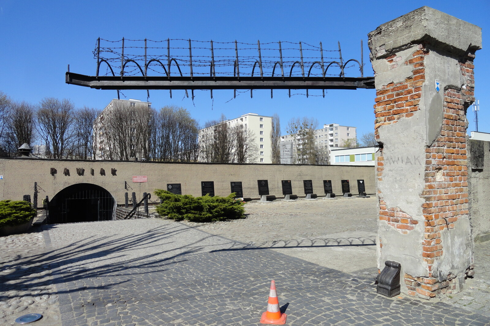 An arch bearing the inscription ‘PAWIAK’ and a bronze tree with plaques against the backdrop of a building.