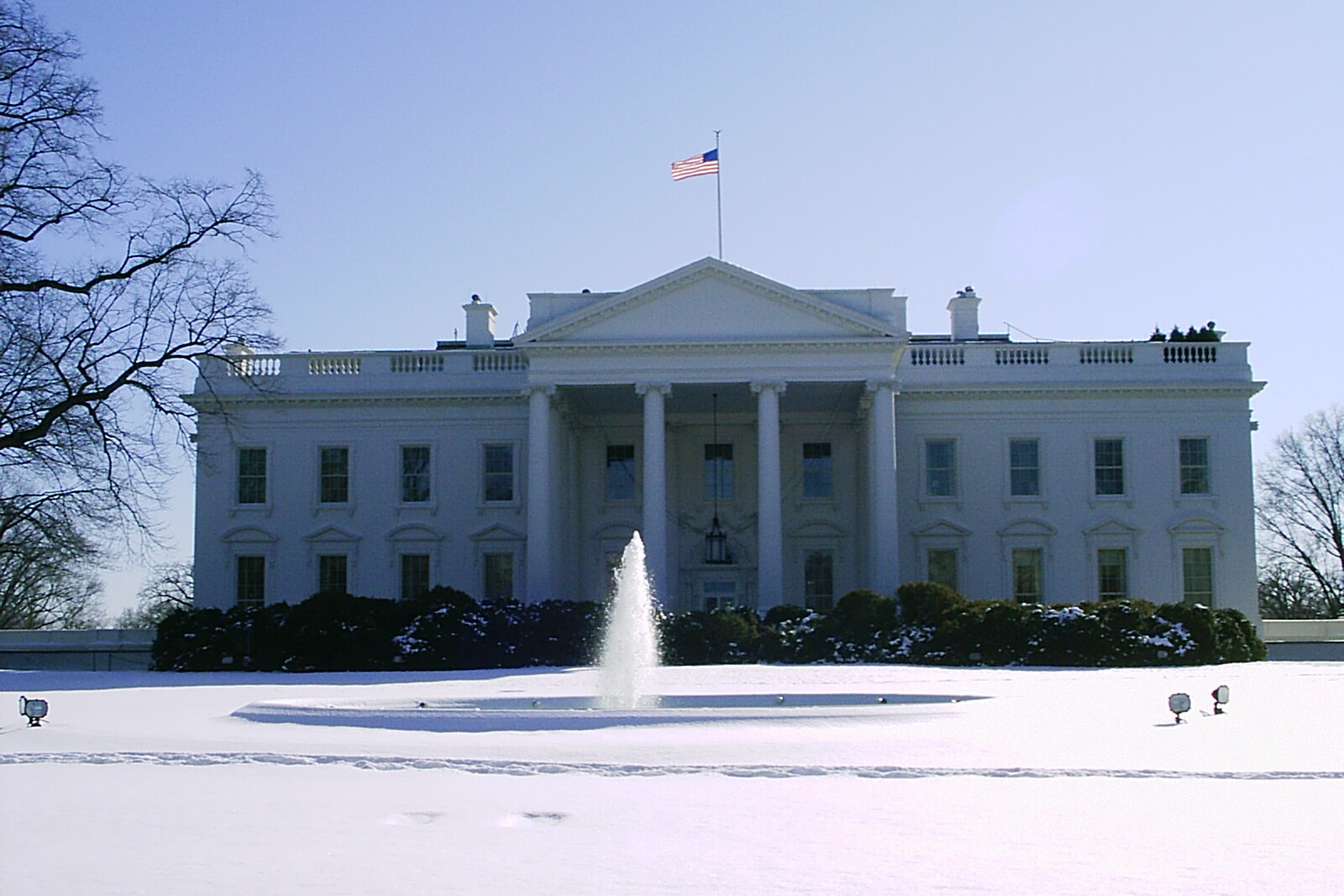The snow-white northern facade of the White House in winter, surrounded by trees and lawn.