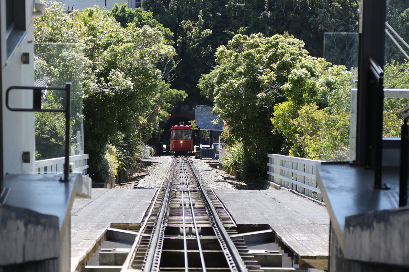A red cable car moves along the tracks against a backdrop of green trees and houses on the hill.